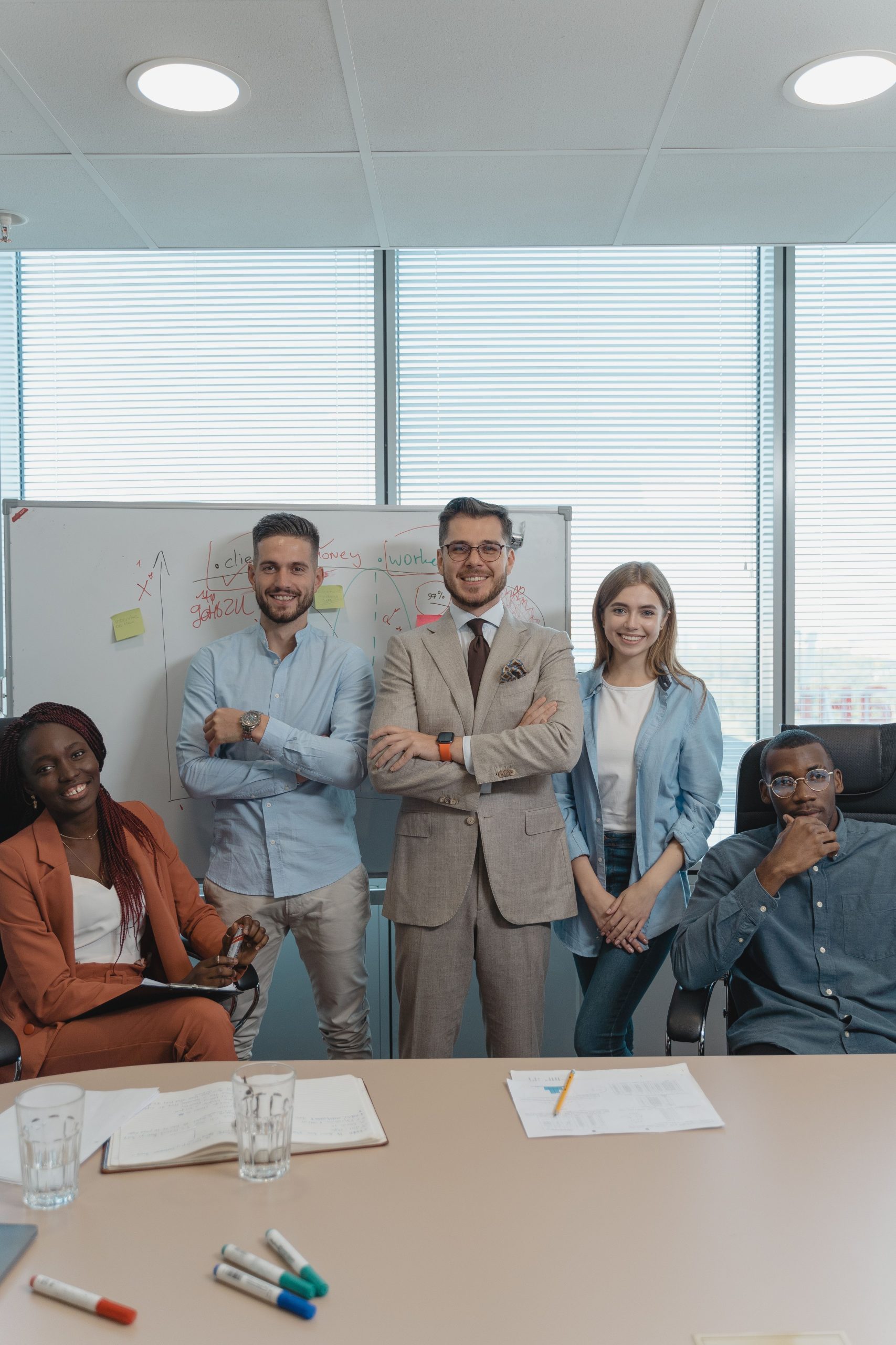 Five coworkers smiling and posing in a bright modern office with a whiteboard and window view.